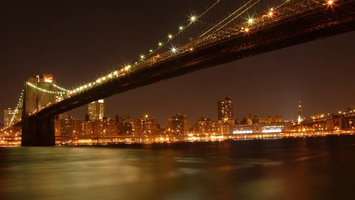 Brooklyn Bridge stretches overhead at night, its lights strung above a glowing Manhattan waterfront.