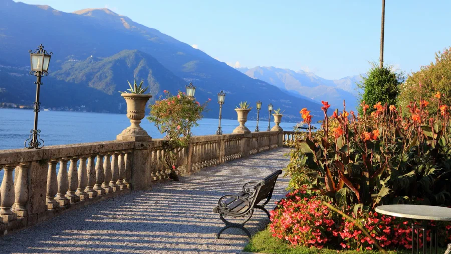 Stone balustrade promenade with lanterns, urns, and flowers curves along a mountain lake in evening sun.