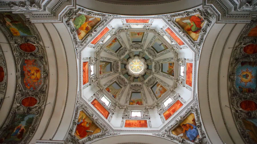 Octagonal baroque cathedral dome seen from below, frescoes and windows ringing a dove at its center.