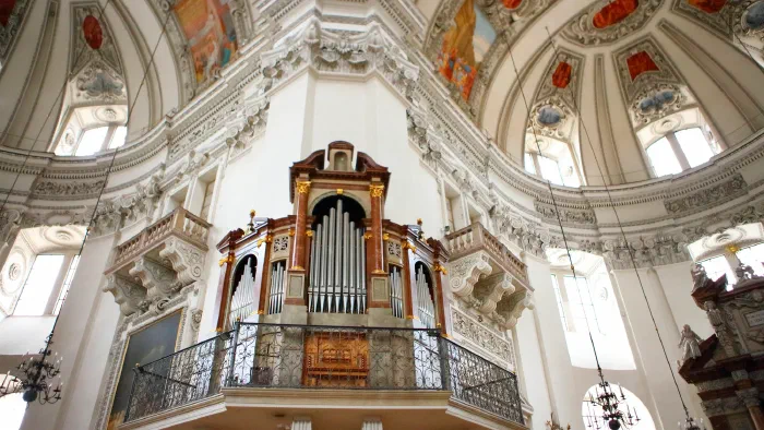 Pipe organ with marble columns and gilded capitals on a balcony beneath white baroque domes.