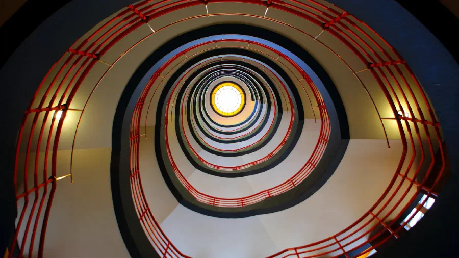 Spiral stairwell with red railings coils upward toward a round stained glass skylight.