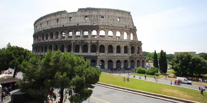 The Colosseum's arched stone tiers rise above green pines and tourists crossing a sunlit street.