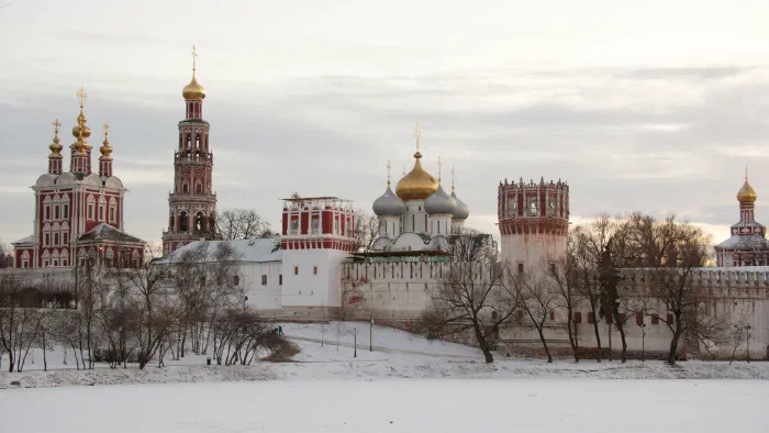 Gold onion domes and red and white towers of a walled Russian monastery rise over snow.
