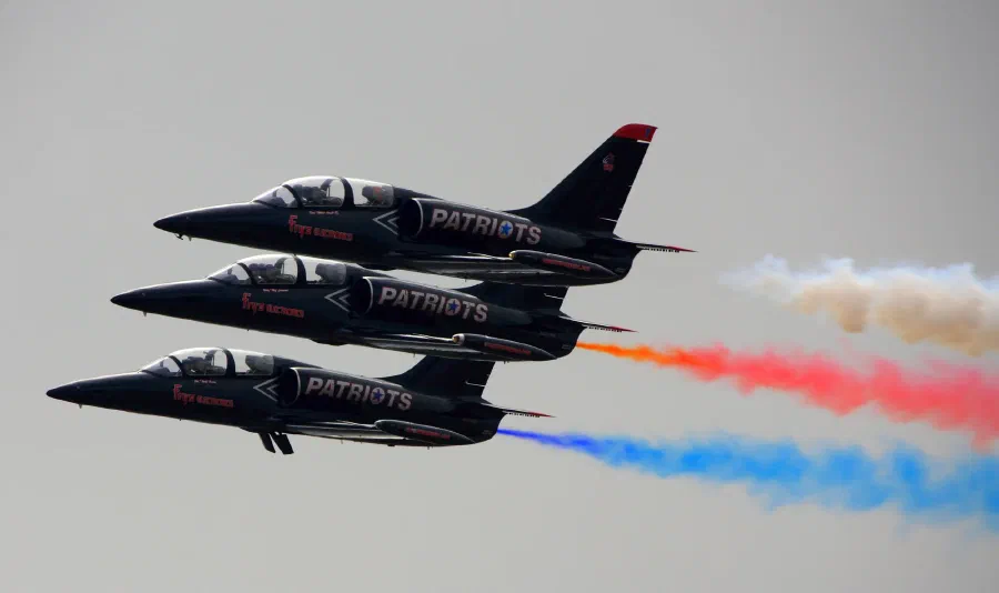 Three black Patriots jets fly in tight stacked formation trailing red, white, and blue smoke.