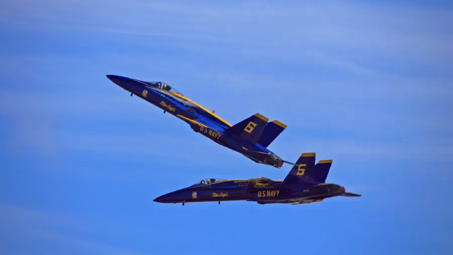 Two Blue Angels jets in blue and gold pass close, one banking skyward above the other.