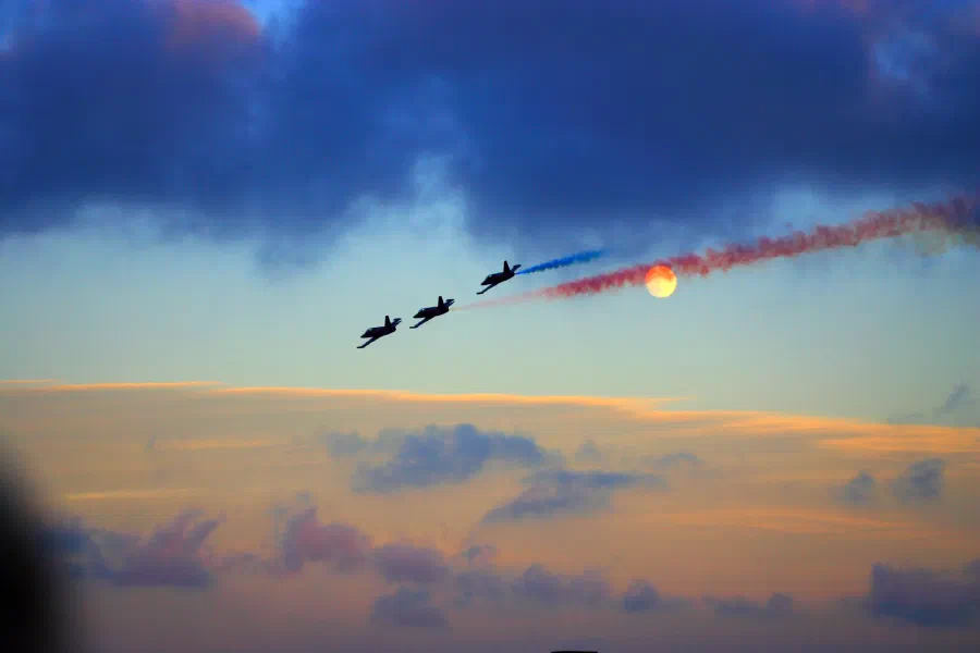 Three jets trail red and blue smoke past a low orange sun in a dusk sky.