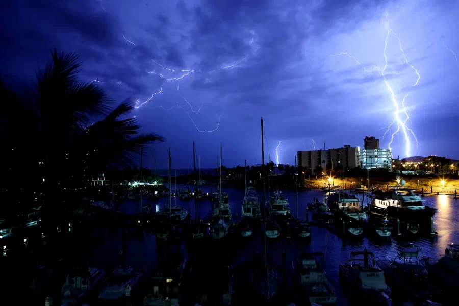 Lightning bolts strike behind waterfront buildings as sailboats rest in a dark marina.