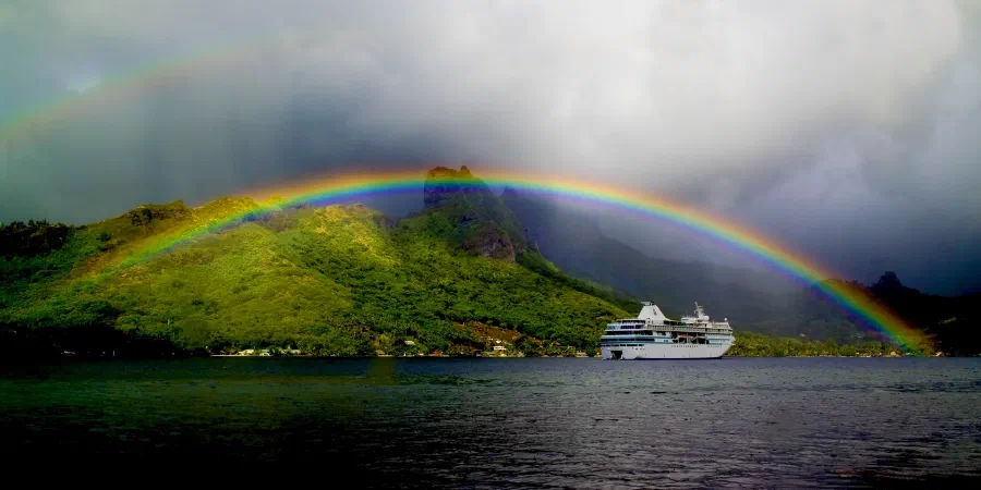 Double rainbow arcs over green volcanic peaks as a white cruise ship anchors beneath it.