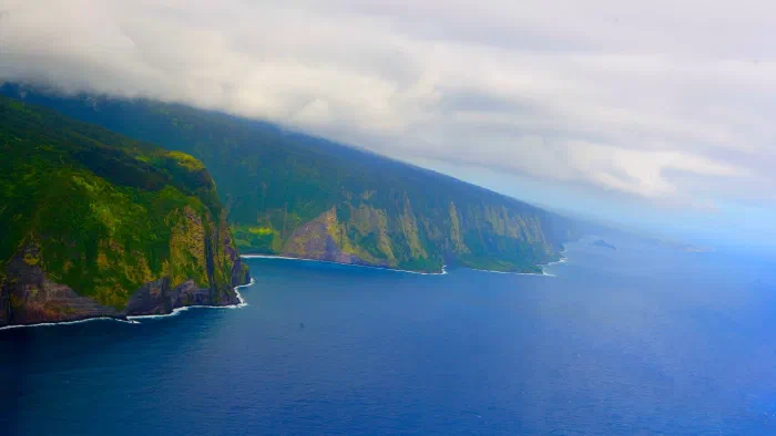 Green sea cliffs laced with waterfalls stretch along a blue coastline under low clouds.