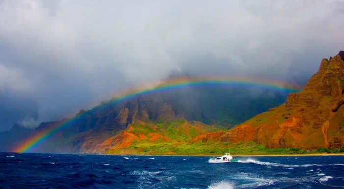 Rainbow arches over rust and green coastal cliffs while a small boat crosses rough blue water.