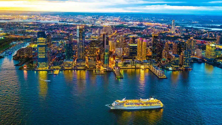 City towers glow at dusk along a river waterfront as a lit cruise ship passes.