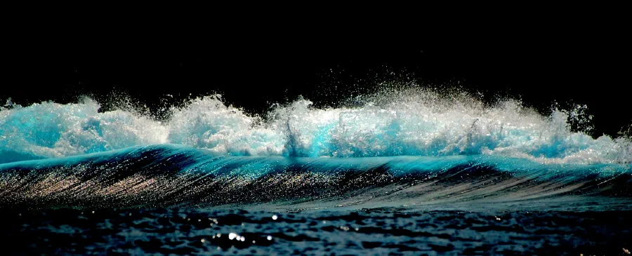 Sunlit wave crest breaks into glittering turquoise spray against a pitch black background.