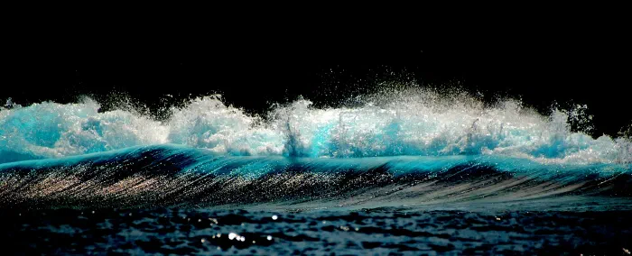 Sunlit wave crest breaks into glittering turquoise spray against a pitch black background.