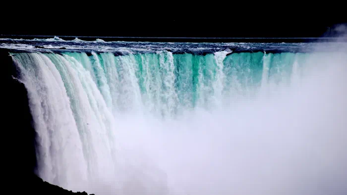 Teal water pours over a broad waterfall brink into a wall of rising white mist.